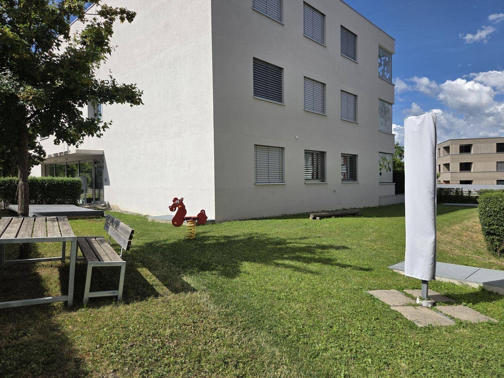 Grüner Gartenbereich mit Holztisch, Bänken und Kinderspielgerät vor einem modernen Gebäude.