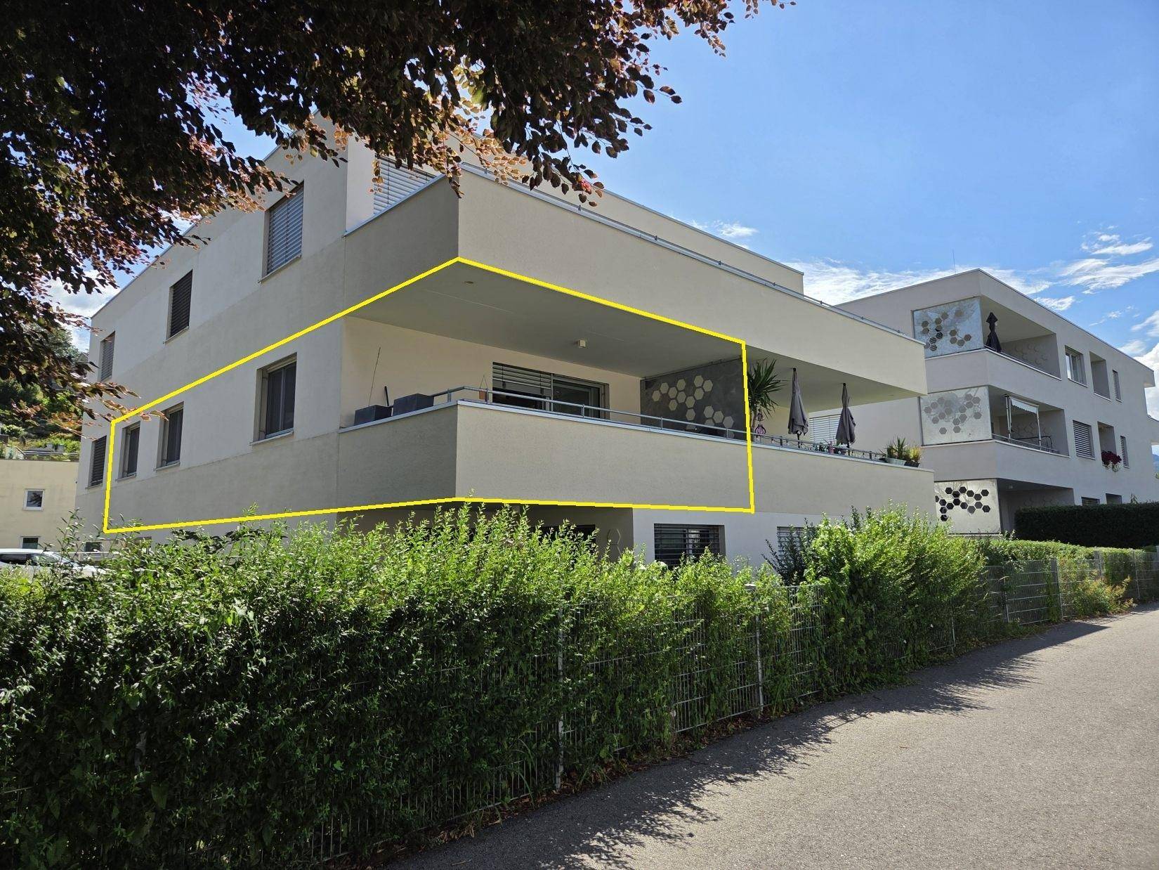 Außenansicht eines modernen Mehrfamilienhauses mit großem Balkon und gepflegter Hecke im Vordergrund.