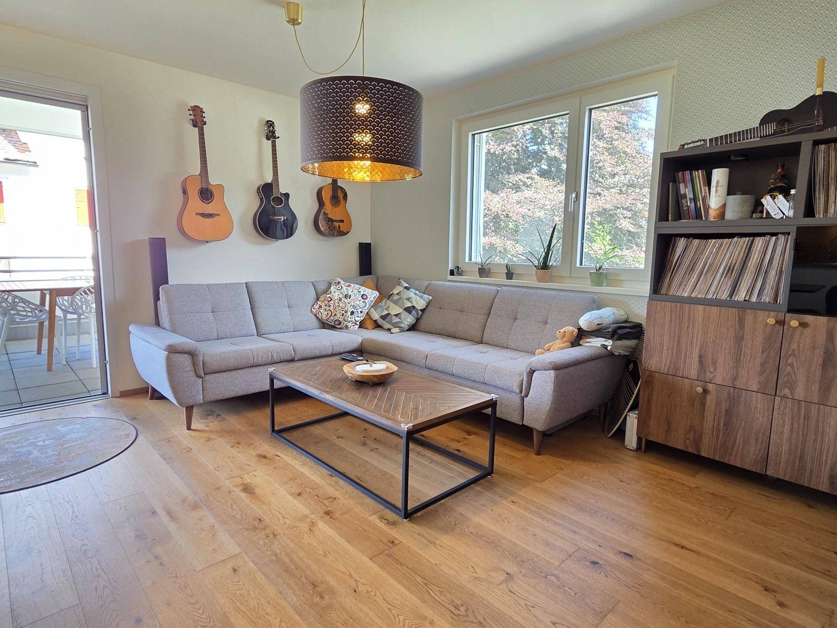 Gemütliches Wohnzimmer mit Ecksofa, Holzboden und direktem Zugang zum sonnigen Balkon.