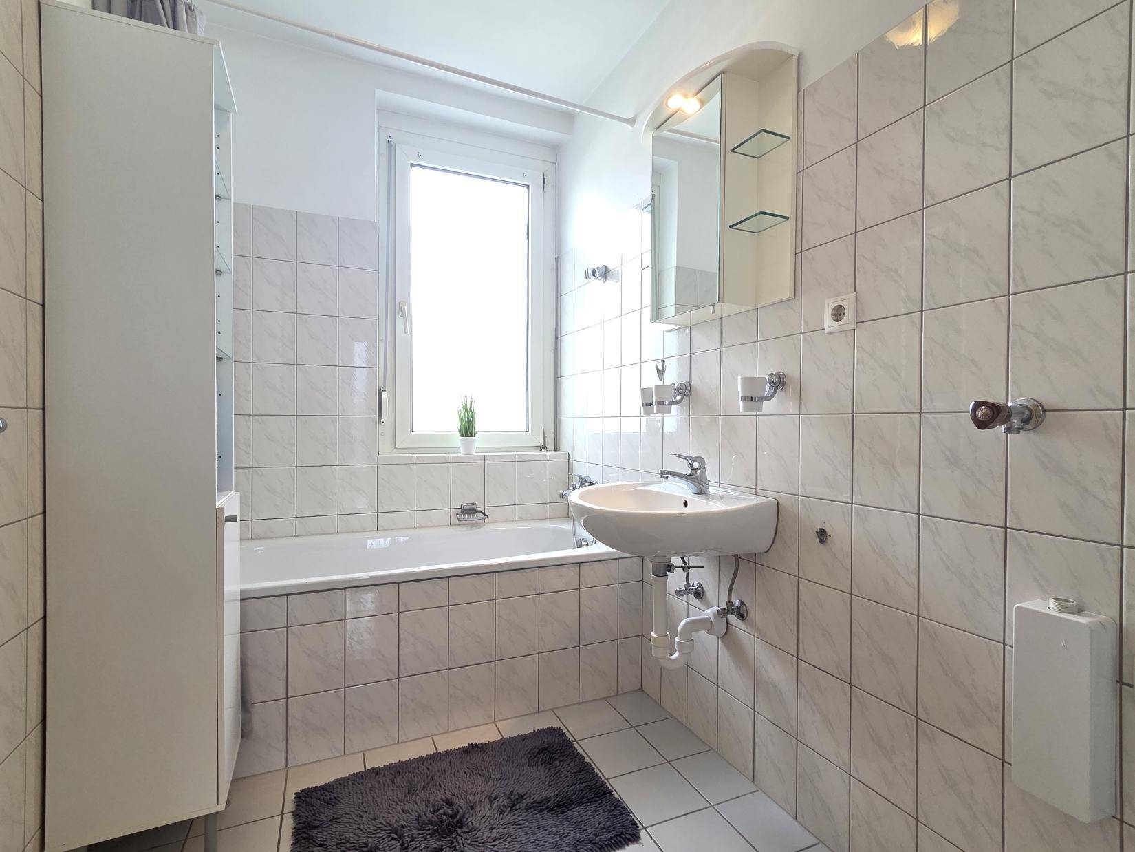 Gefliestes Badezimmer mit Badewanne, Waschbecken und Fenster für natürliche Belüftung.