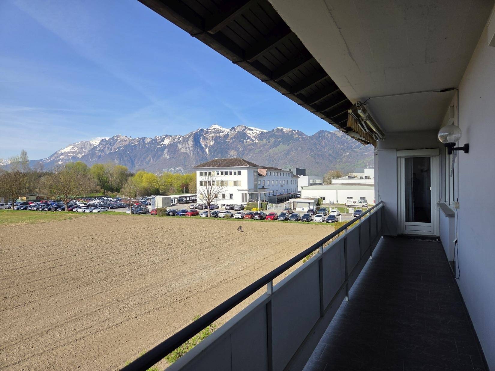 Balkon mit weitem Blick über Felder, Parkplatz und die beeindruckende Berglandschaft.