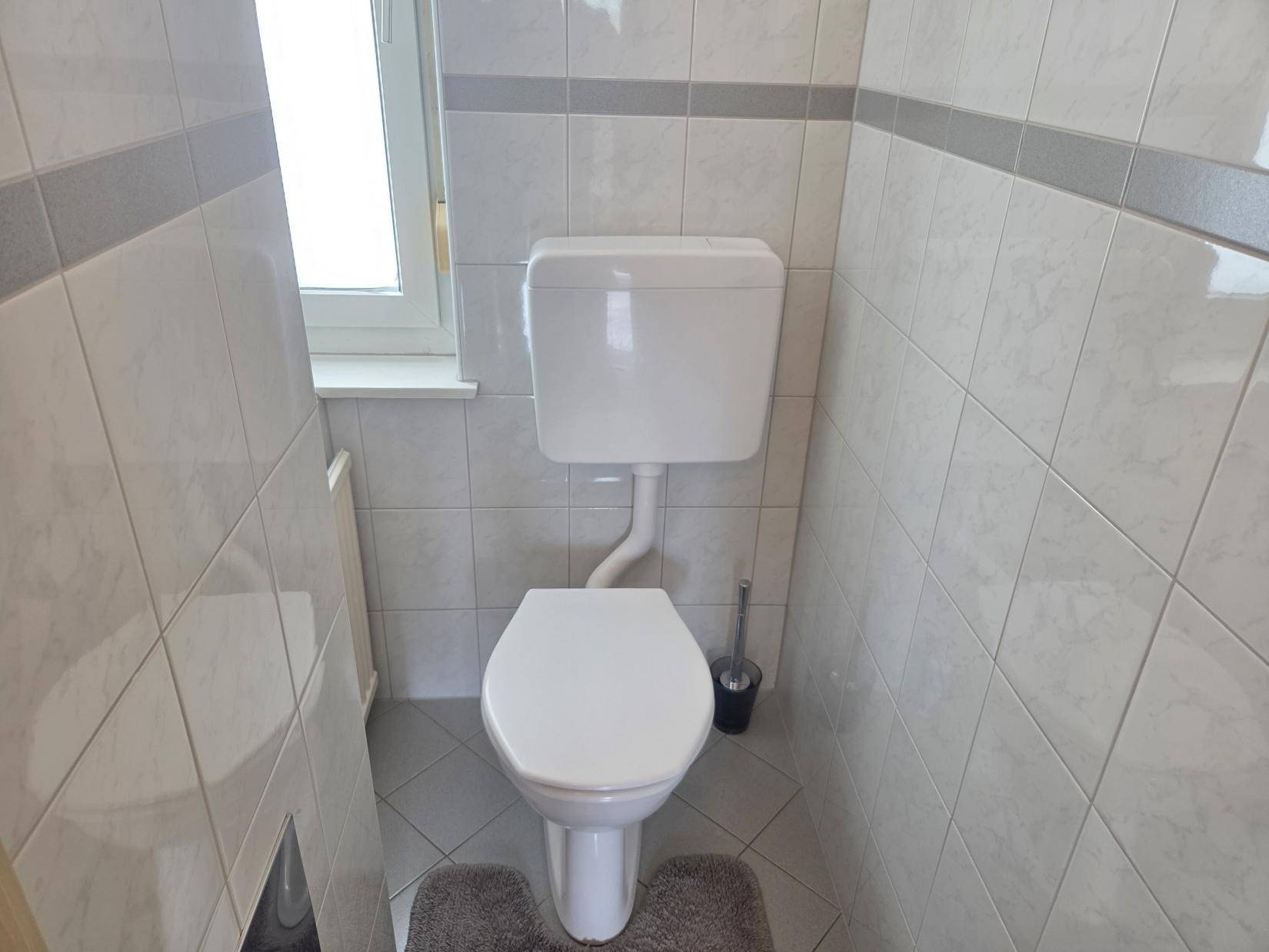 Separates WC mit Toilette, hellen Fliesen und kleinem Fenster für Belüftung.