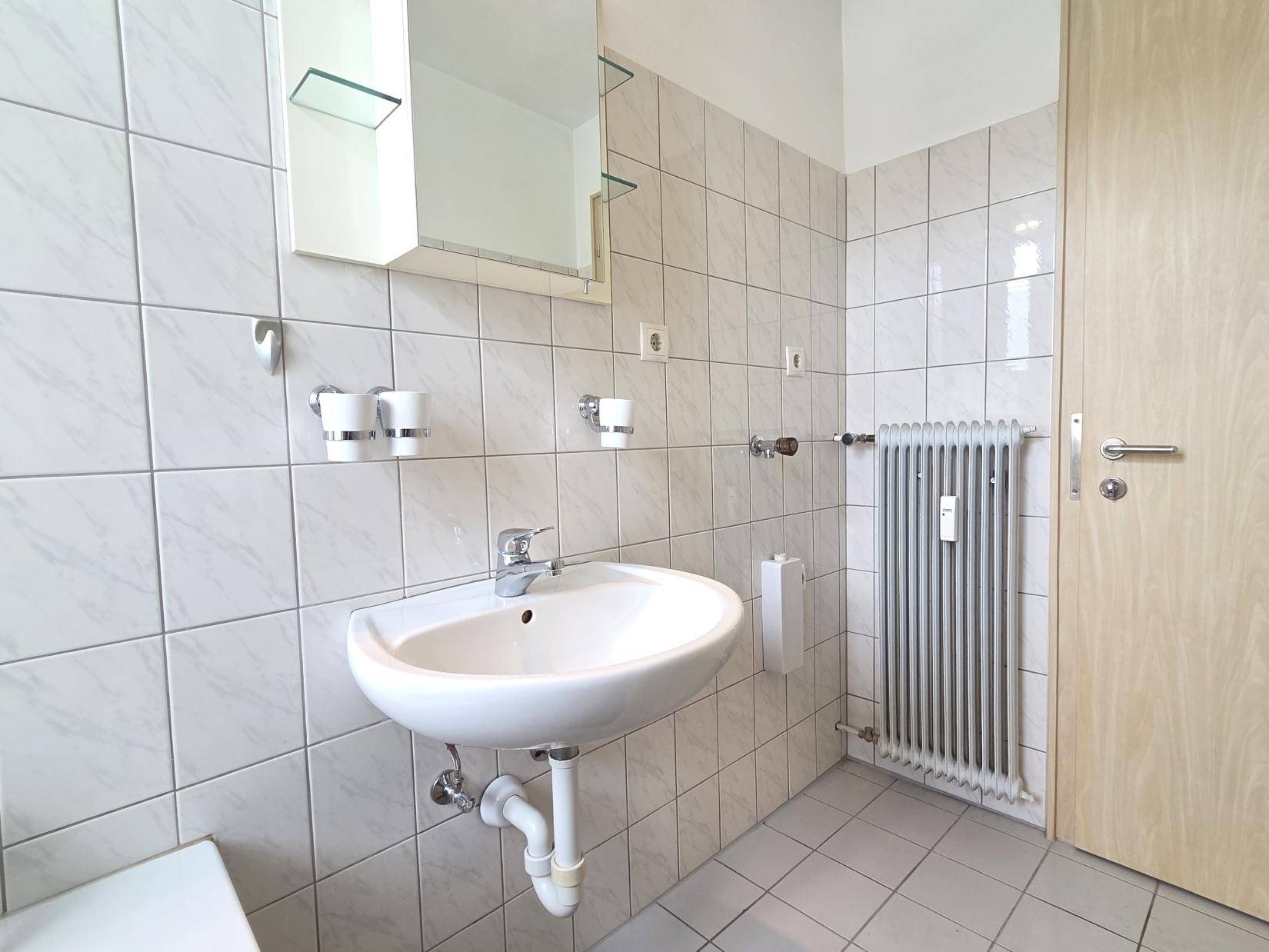Badezimmer mit Waschbecken, Spiegelschrank und Heizkörper, alles in hellen Fliesen gehalten.