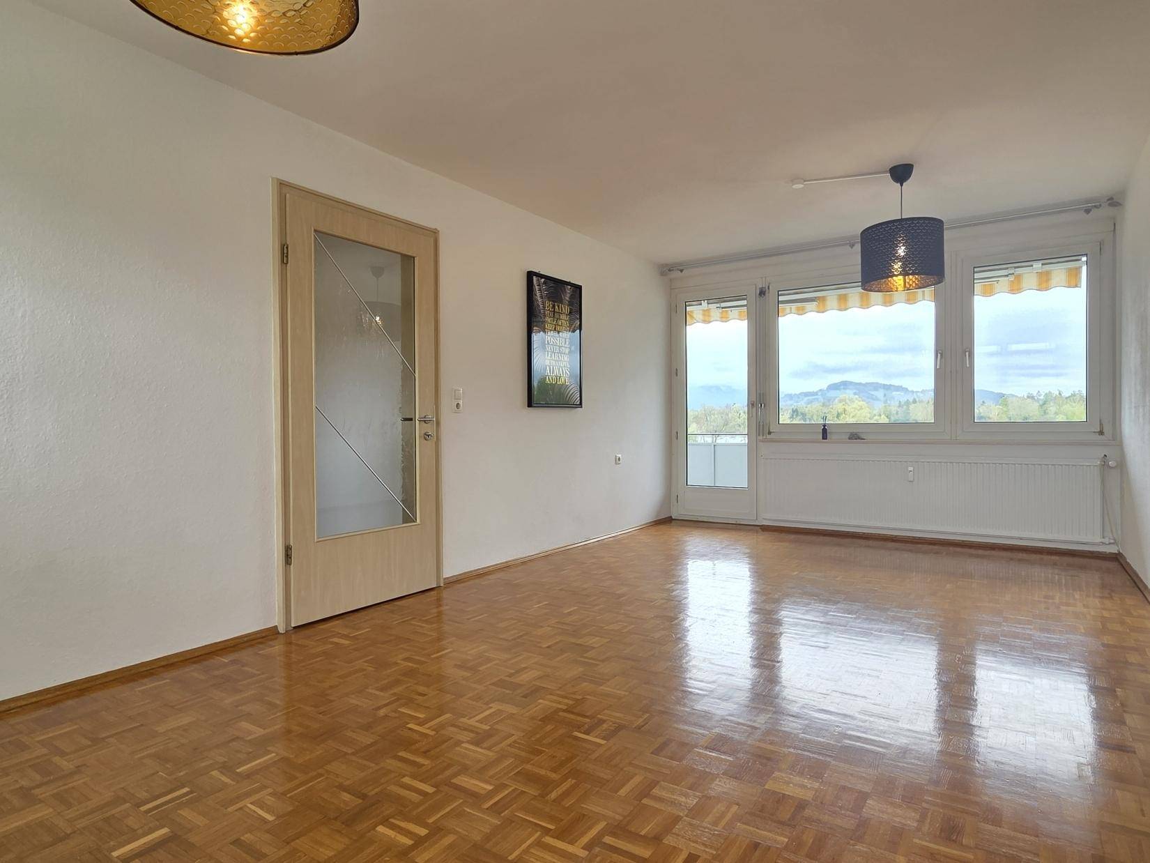 Geräumiges Wohnzimmer mit Parkettboden, viel Tageslicht und Bergblick durch die Fenster.
