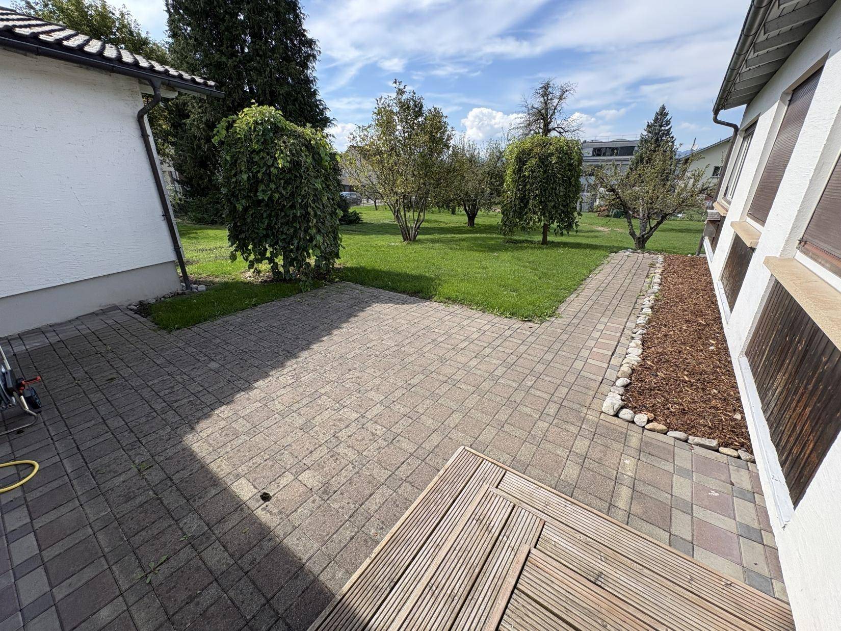Gepflasterter Terrassenbereich mit Holzdeck und Zugang zum grünen Garten.