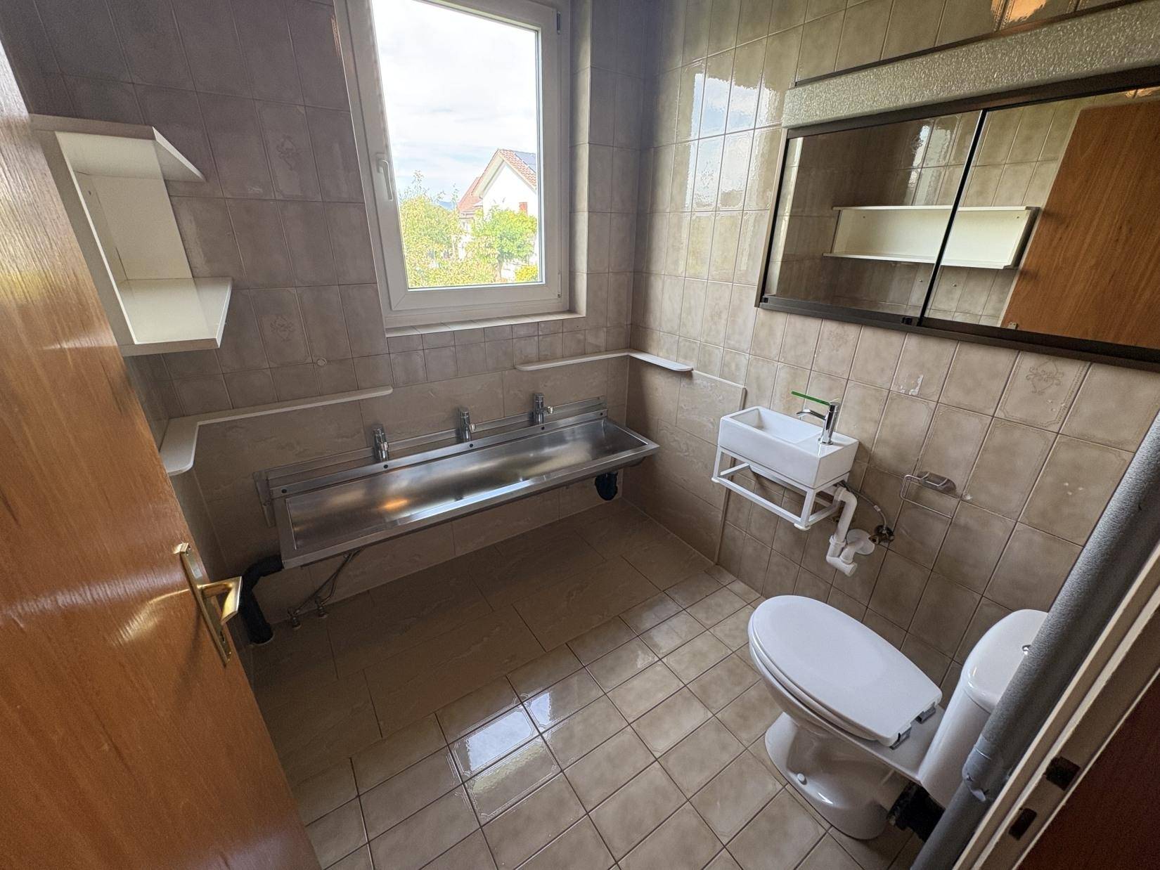 Gefliestes Badezimmer mit Toilette, mehreren Waschbecken und Fenster.