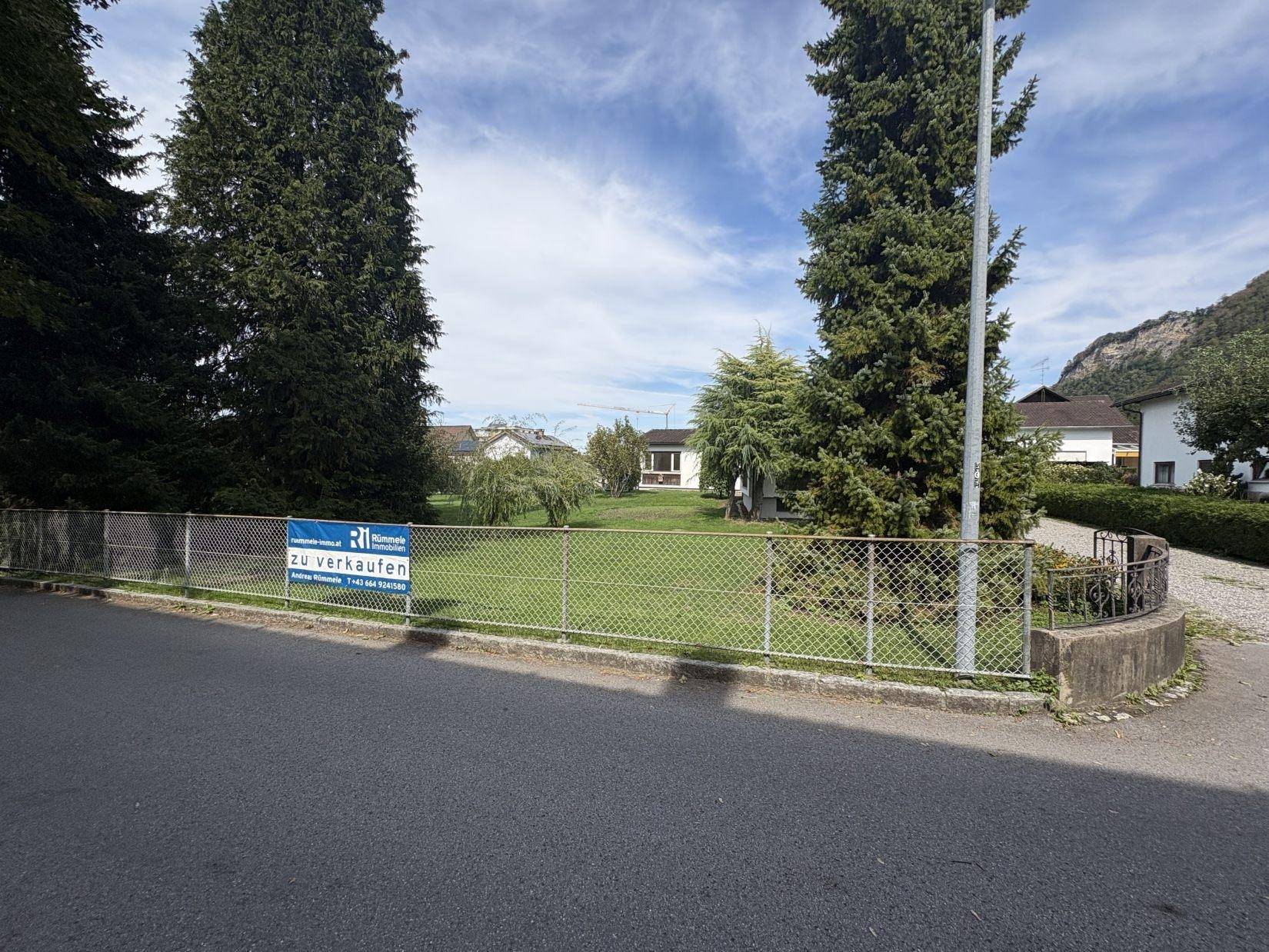 Grundstück mit Einfamilienhaus und 'Zu verkaufen'-Schild am Zaun, umgeben von Natur und Bergen.