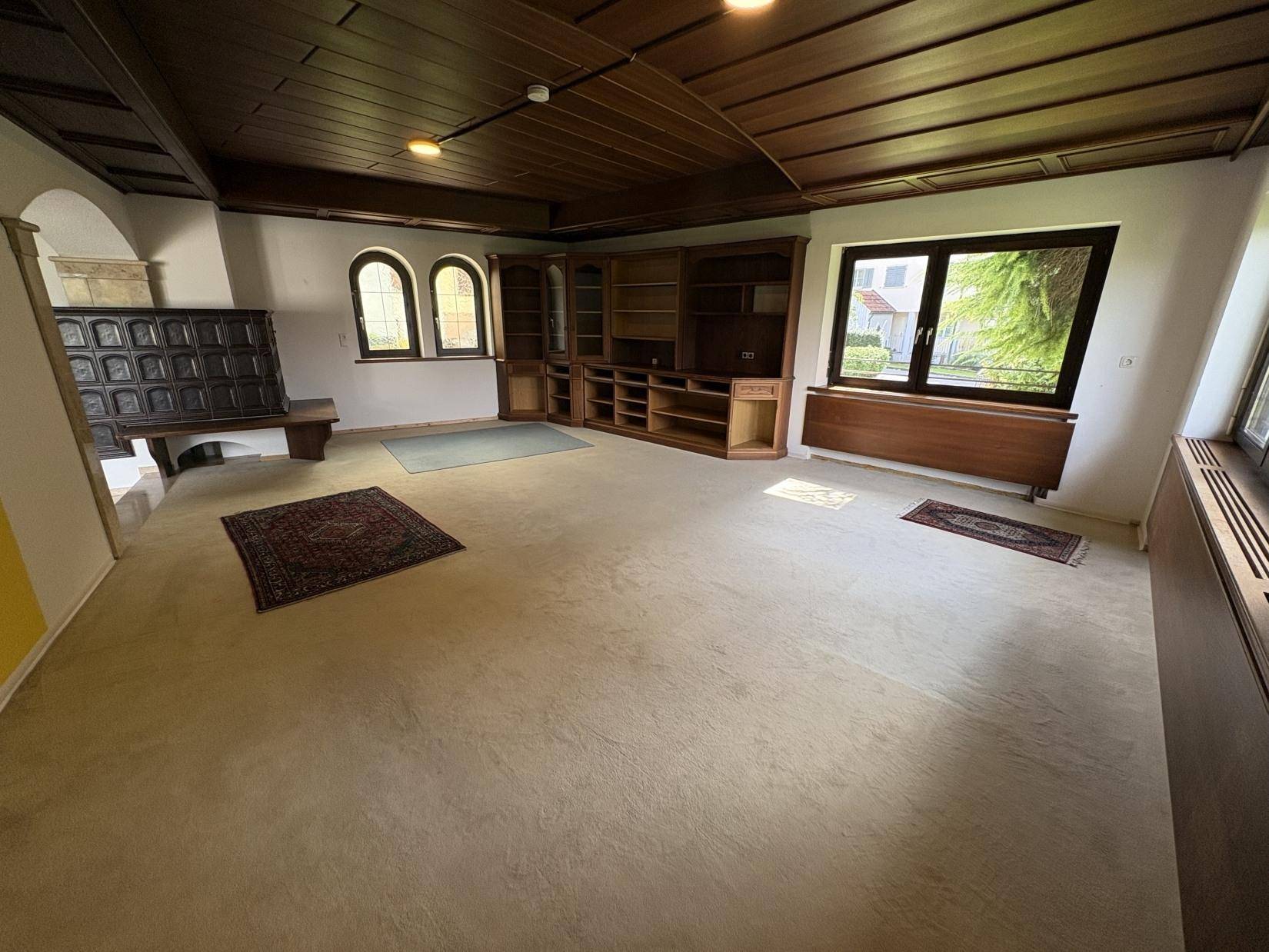 Geräumiges Wohnzimmer mit Teppichboden, Holzkassettendecke und traditionellem Kachelofen.