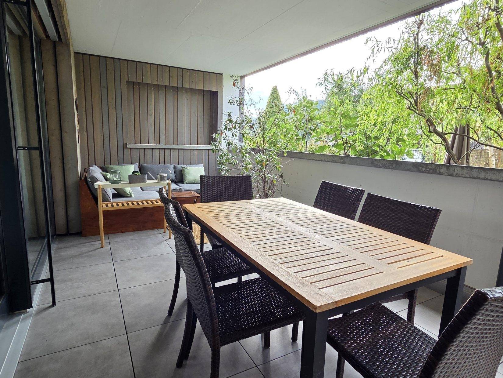 Überdachter Balkon mit Esstisch und Lounge-Bereich, umgeben von Grün.