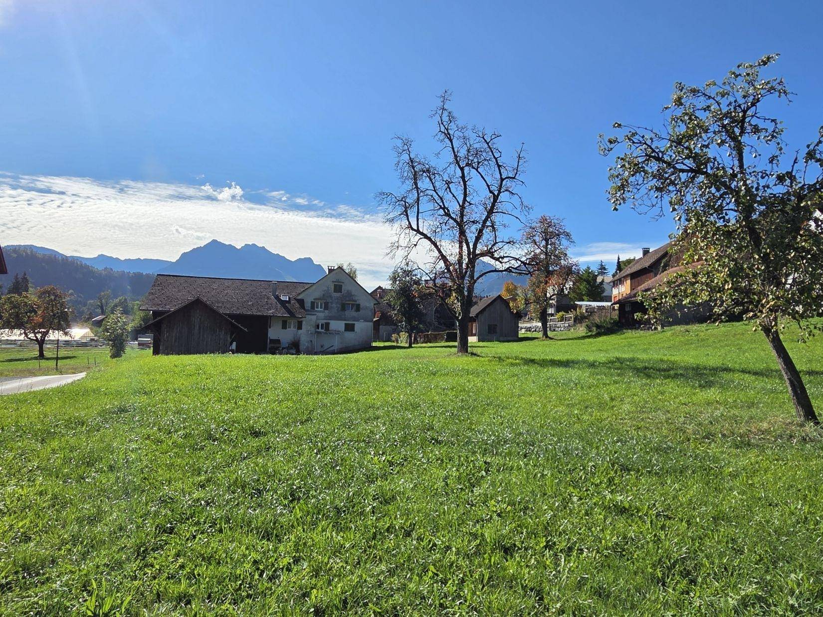 Weitläufige grüne Wiese mit vereinzelten Bäumen und traditionellen Gebäuden im Hintergrund, umgeben von Bergen.