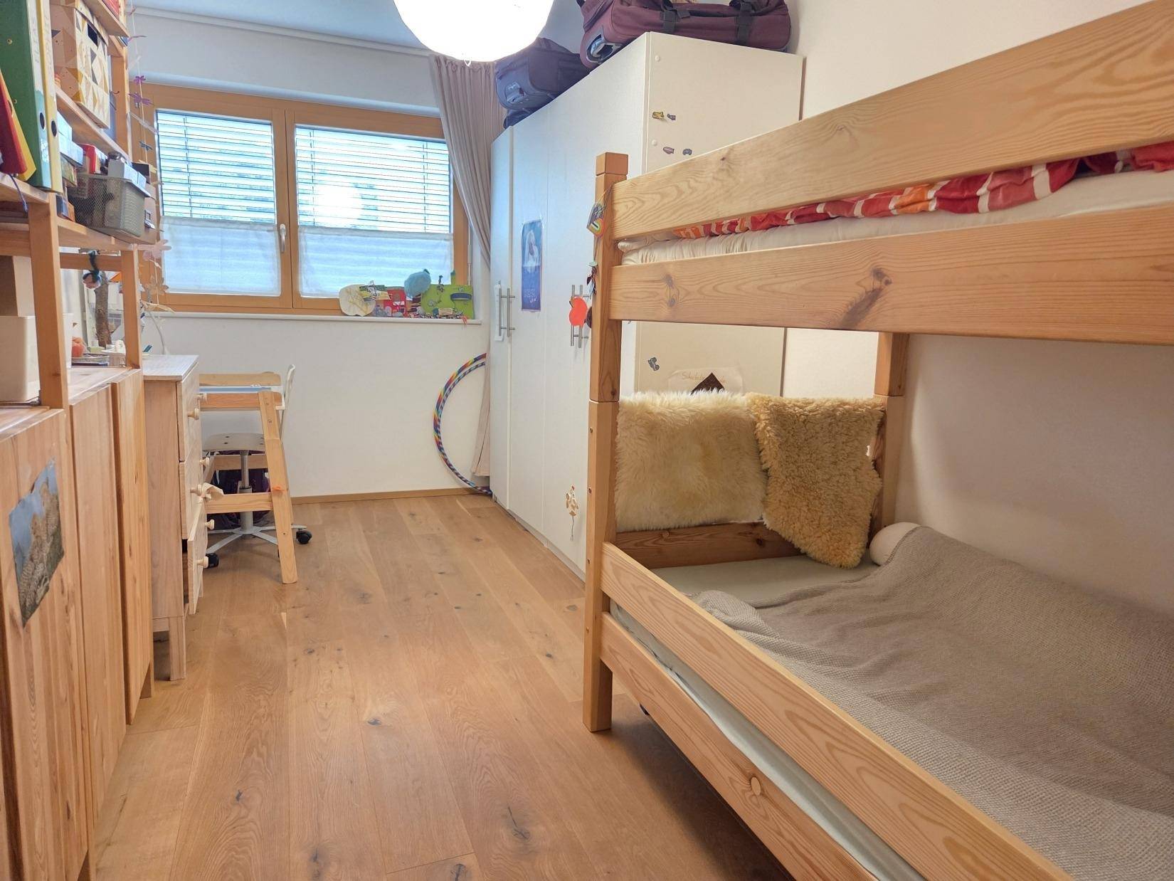 Helles Kinderzimmer mit Etagenbett, Schreibtisch und Fenster mit Jalousien.