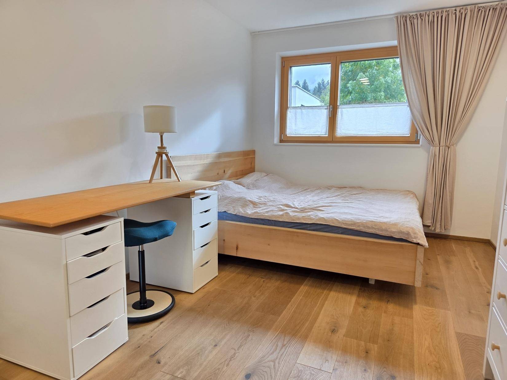 Schlafzimmer mit Holzbett, Schreibtisch und großem Fenster für viel Tageslicht.