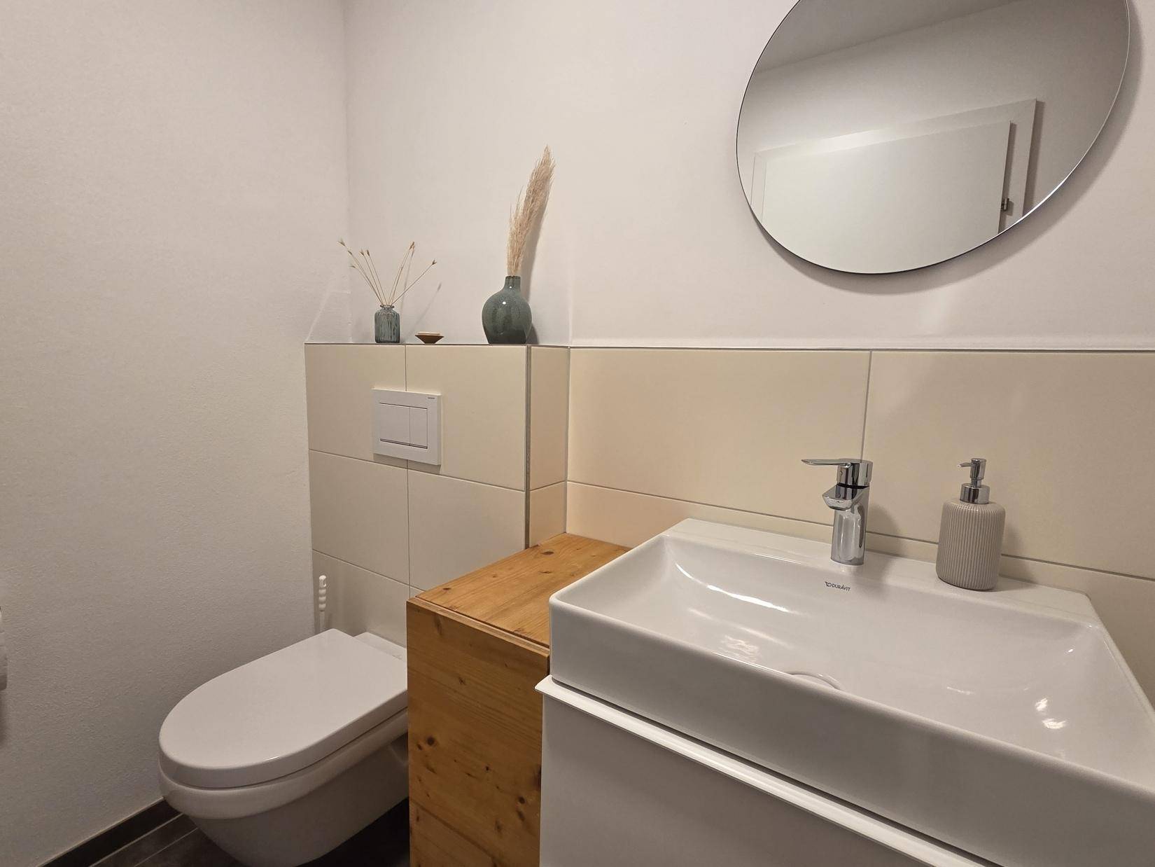 Gäste-WC mit modernem Waschbecken, Wand-WC und rundem Spiegel.