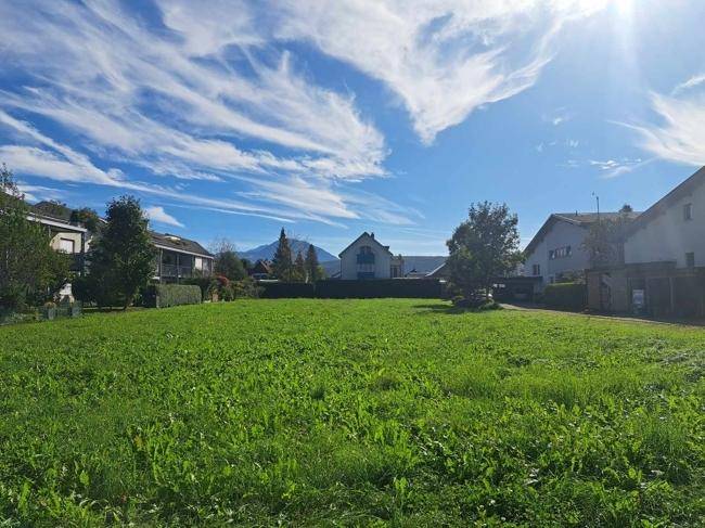 Weitläufige Grünfläche mit Blick auf umliegende Häuser und Berge unter strahlend blauem Himmel.