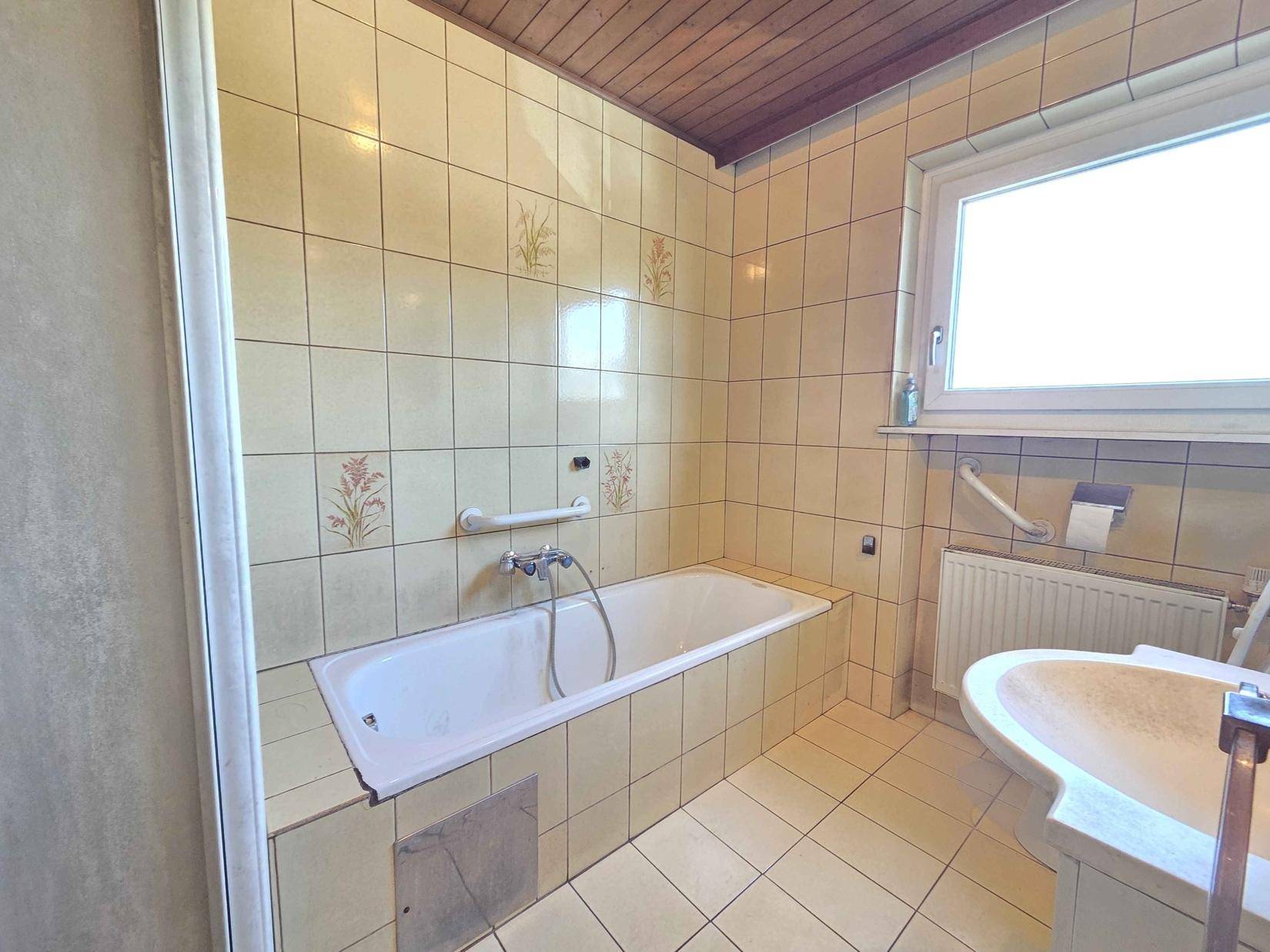 Badezimmer im 80er-Jahre-Stil mit Badewanne, Dusche, Waschbecken und Haltegriffen.