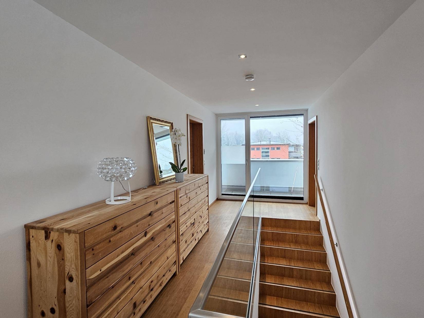 Lichtdurchfluteter Flur mit Holztreppe, Glasgeländer und Zugang zu einem Balkon.