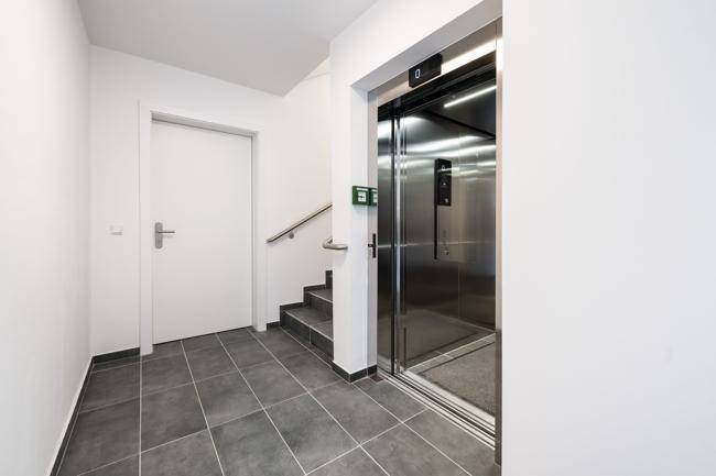 Moderner Eingangsbereich mit Aufzug, Treppe und Fliesenboden.