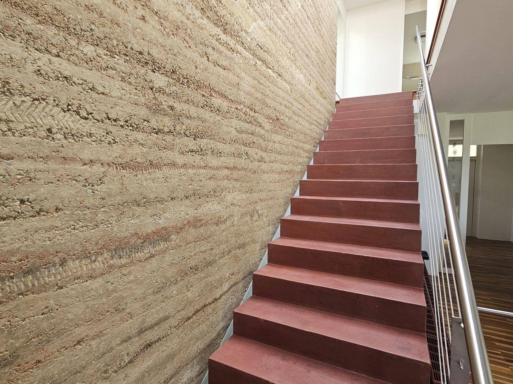 Moderne Treppe mit roten Stufen und einer Wand aus gestampftem Lehm, die zum Obergeschoss führt.