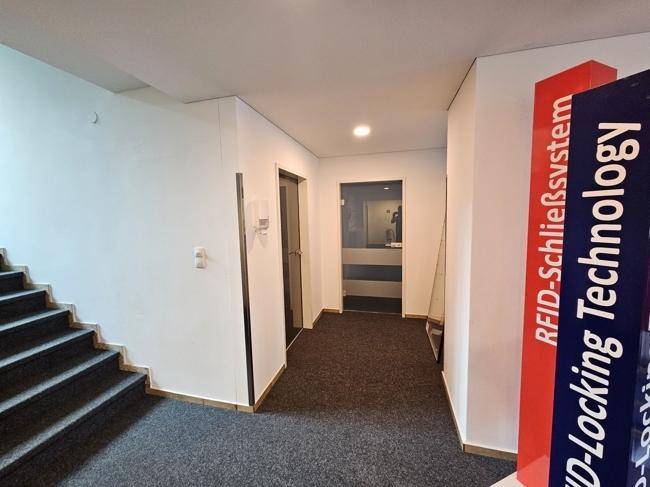 Moderner Flur mit Teppichboden, weißen Wänden und einer Glastür, die zu weiteren Räumen führt.