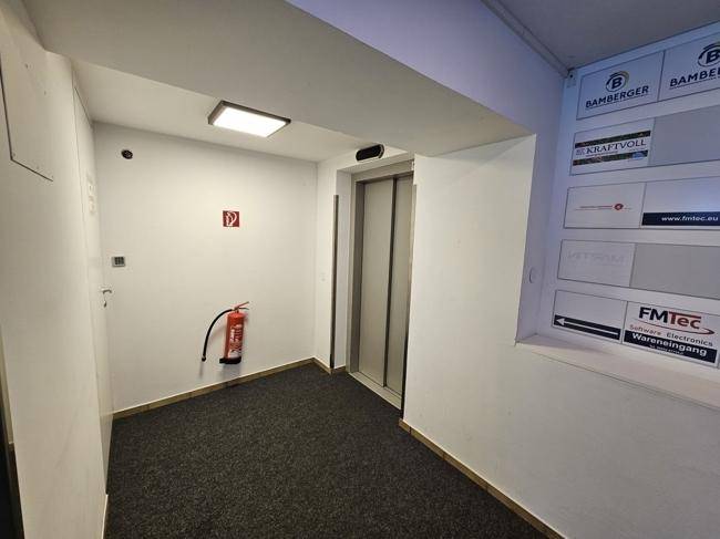 Aufzugsbereich mit Teppichboden, weißen Wänden und einem Feuerlöscher, gut beleuchtet.