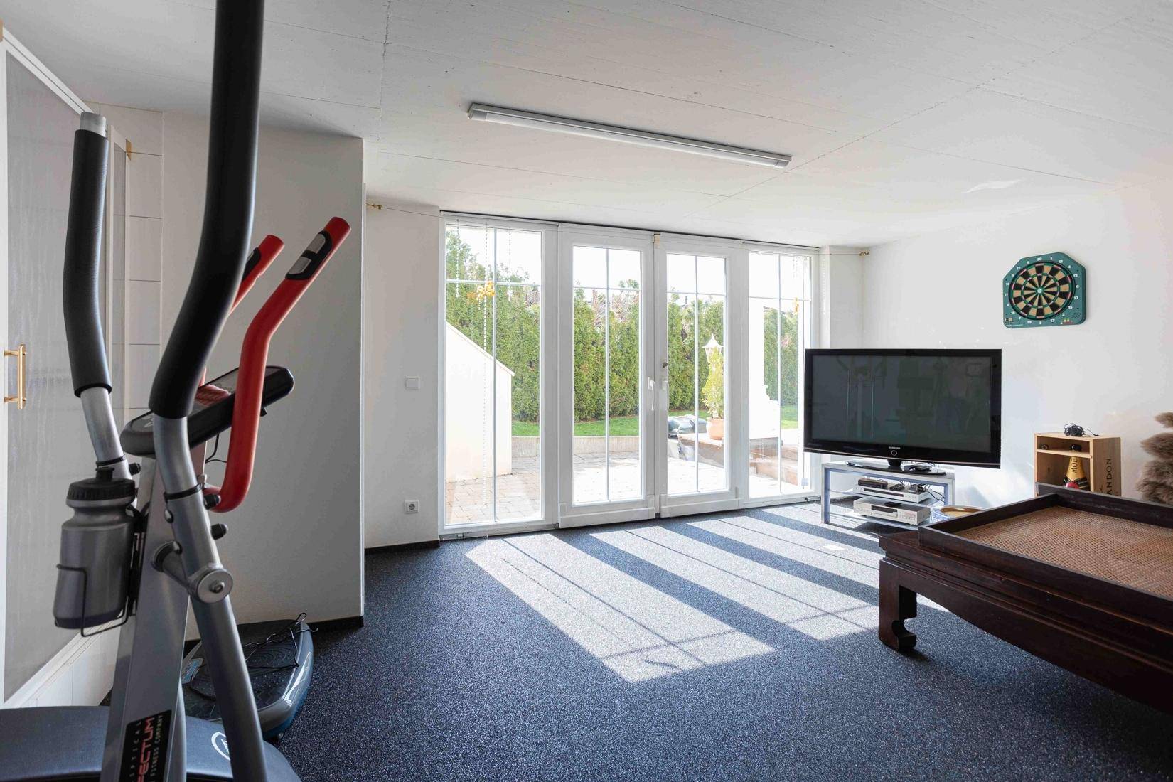Heller Fitnessraum mit großen Fenstern, Fernseher und Zugang zum Garten.
