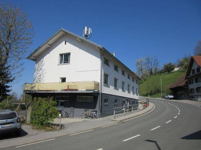 Weißes Gebäude mit dunklem Sockel und Balkon an einer kurvigen Straße in ländlicher Umgebung.