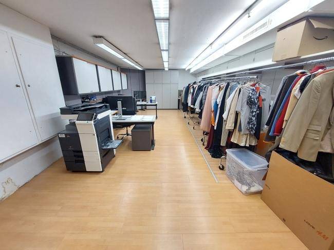 Hinterraum mit Arbeitsplatz, Drucker und Kleiderständern, genutzt als Büro und Lagerbereich.