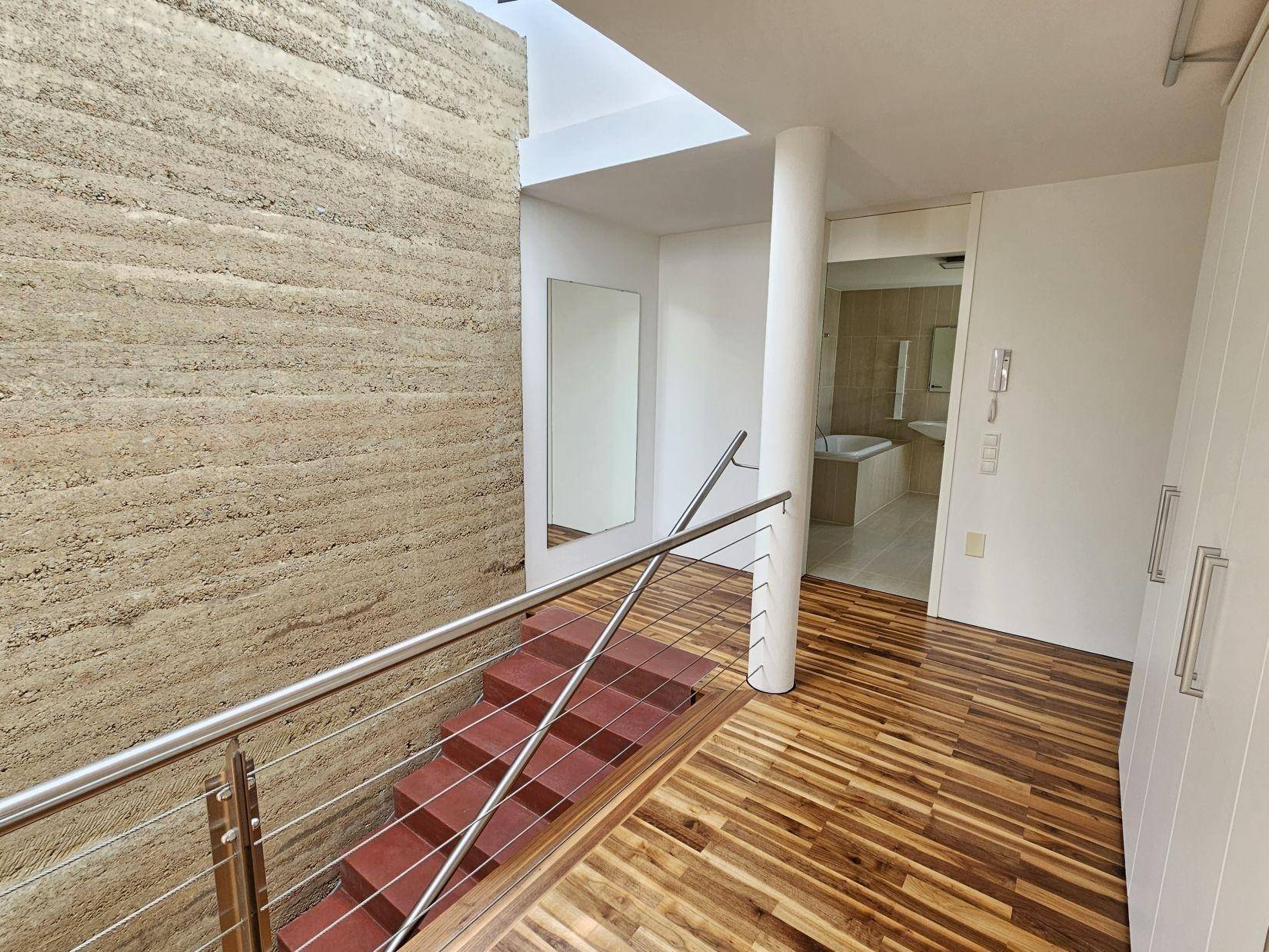 Heller Flur im Obergeschoss mit Holzparkett, Edelstahlgeländer und Blick auf das Badezimmer.