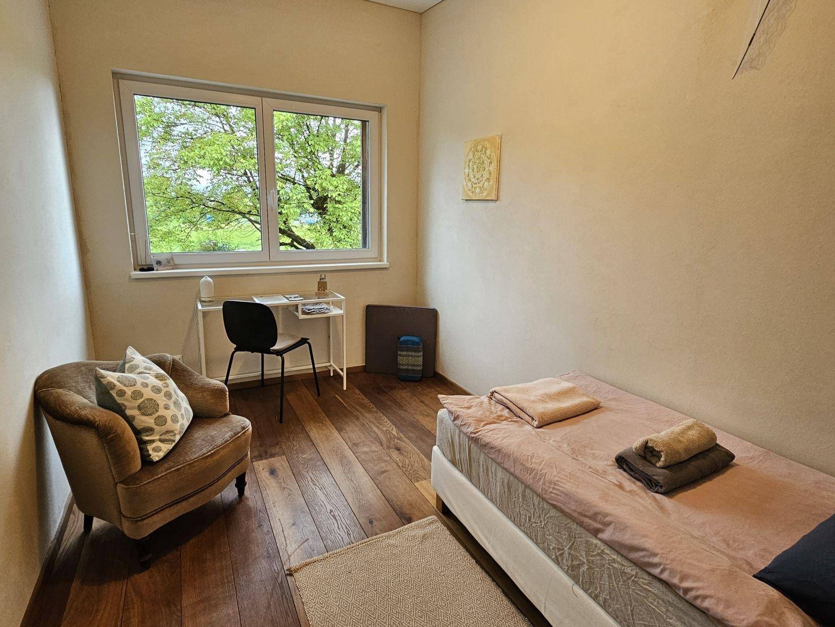 Gemütliches Schlafzimmer mit Einzelbett, Sessel und Fenster, das viel Tageslicht hereinlässt.