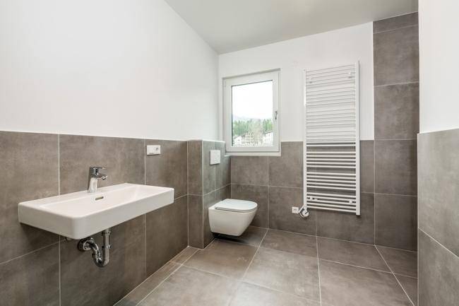 Modernes Badezimmer mit grauen Fliesen, wandhängendem WC, Waschbecken und Fenster.