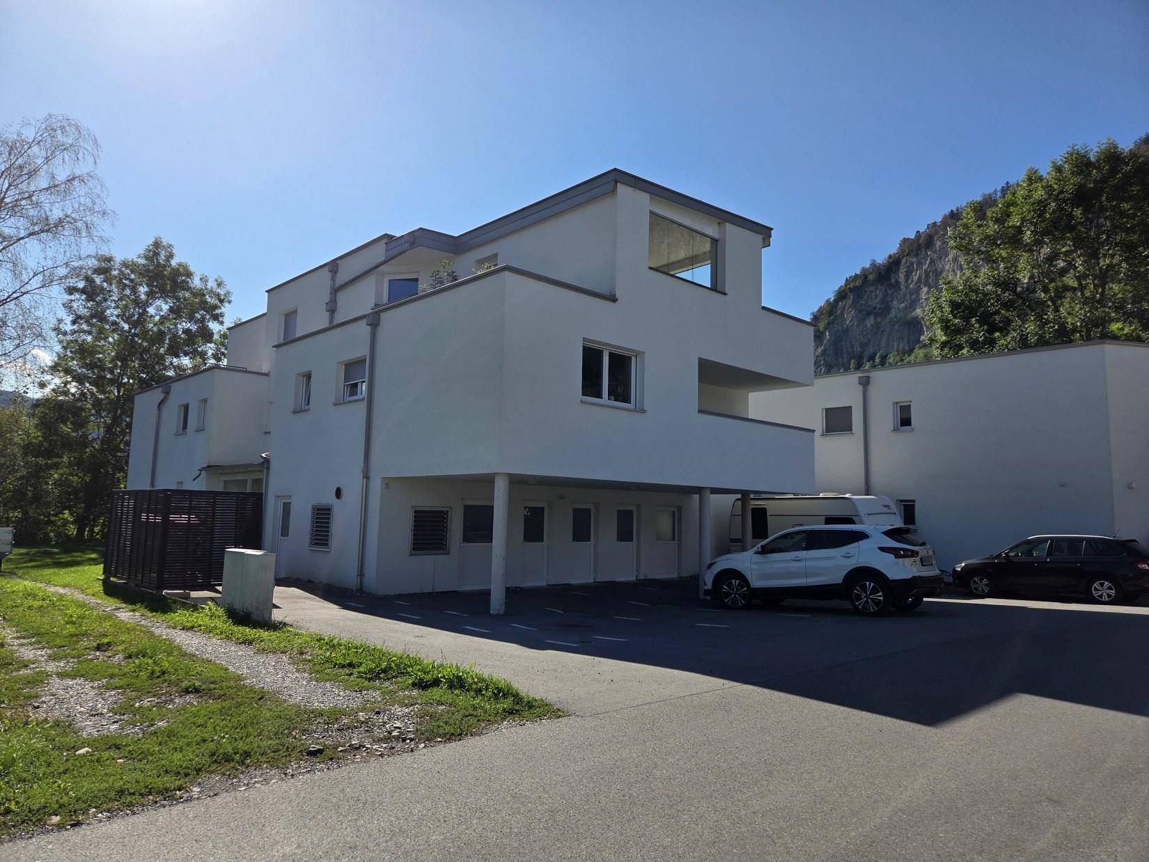 Moderne Gebäudefassade mit weißem Putz, Flachdach und Parkplätzen vor einer Bergkulisse.