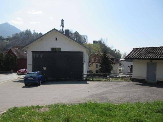 Gebäuderückseite mit großem schwarzen Anbau und Parkplatz vor einer bergigen Landschaft.