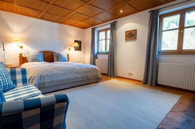 Gemütliches Schlafzimmer mit Doppelbett, kariertem Sessel und Fenstern, die den Raum mit Licht füllen.
