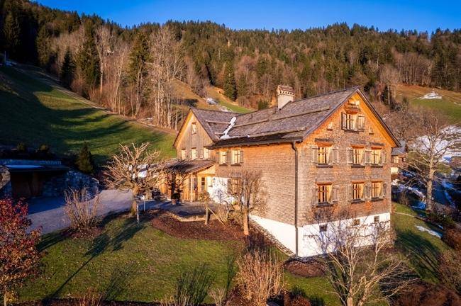 Das traditionelle Holzhaus in sonniger Lage, umgeben von grünen Hängen und Bäumen.