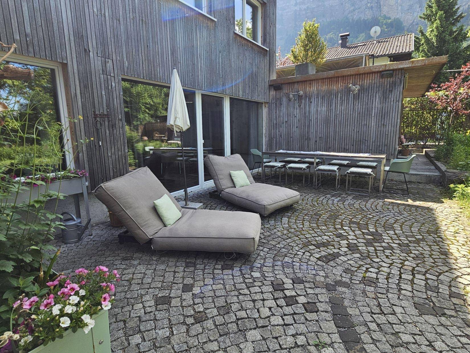 Gepflasterte Terrasse mit Liegestühlen und einem Esstisch im Freien, umgeben von Grün.