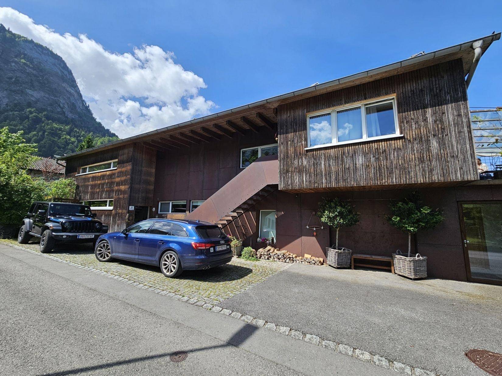 Modernes Gebäude mit Holzfassade, Außentreppe und Parkplätzen vor einer beeindruckenden Bergkulisse.