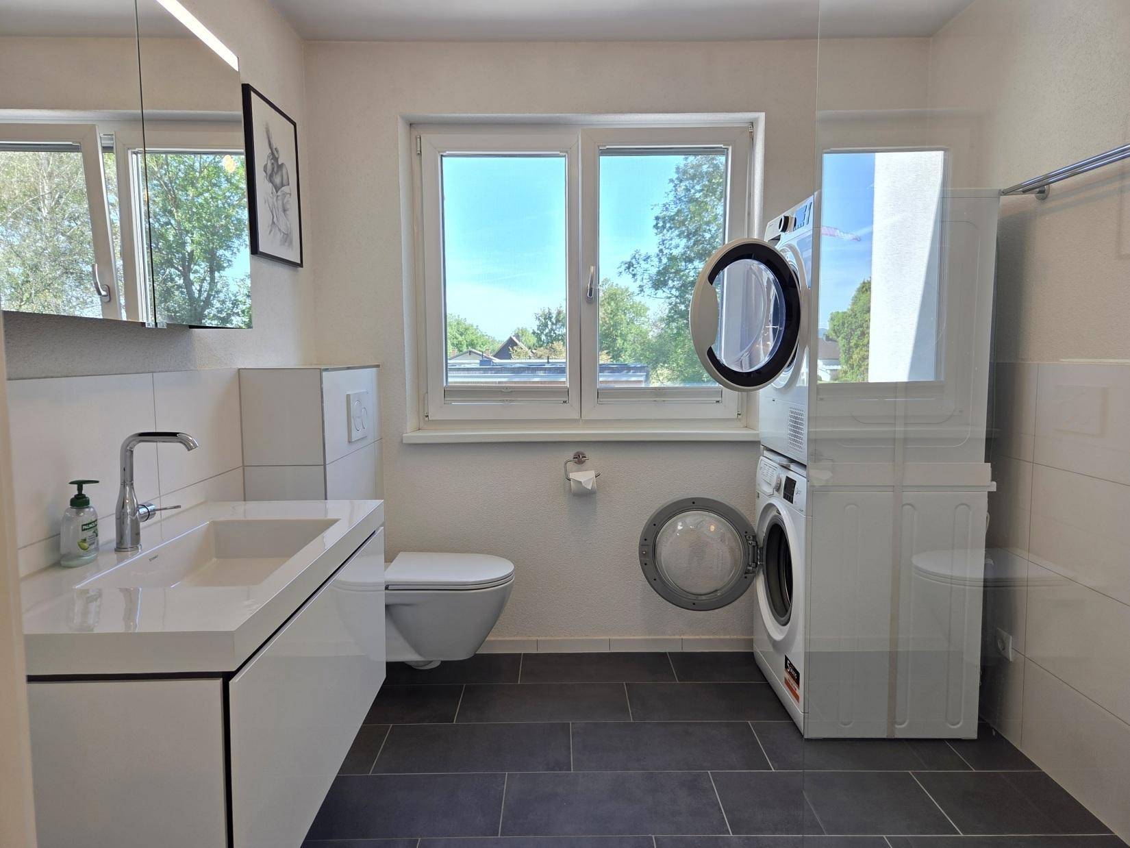 Modernes Badezimmer mit dunklen Fliesen, schwebendem Waschtisch, Wand-WC und Waschmaschine mit Trockner.