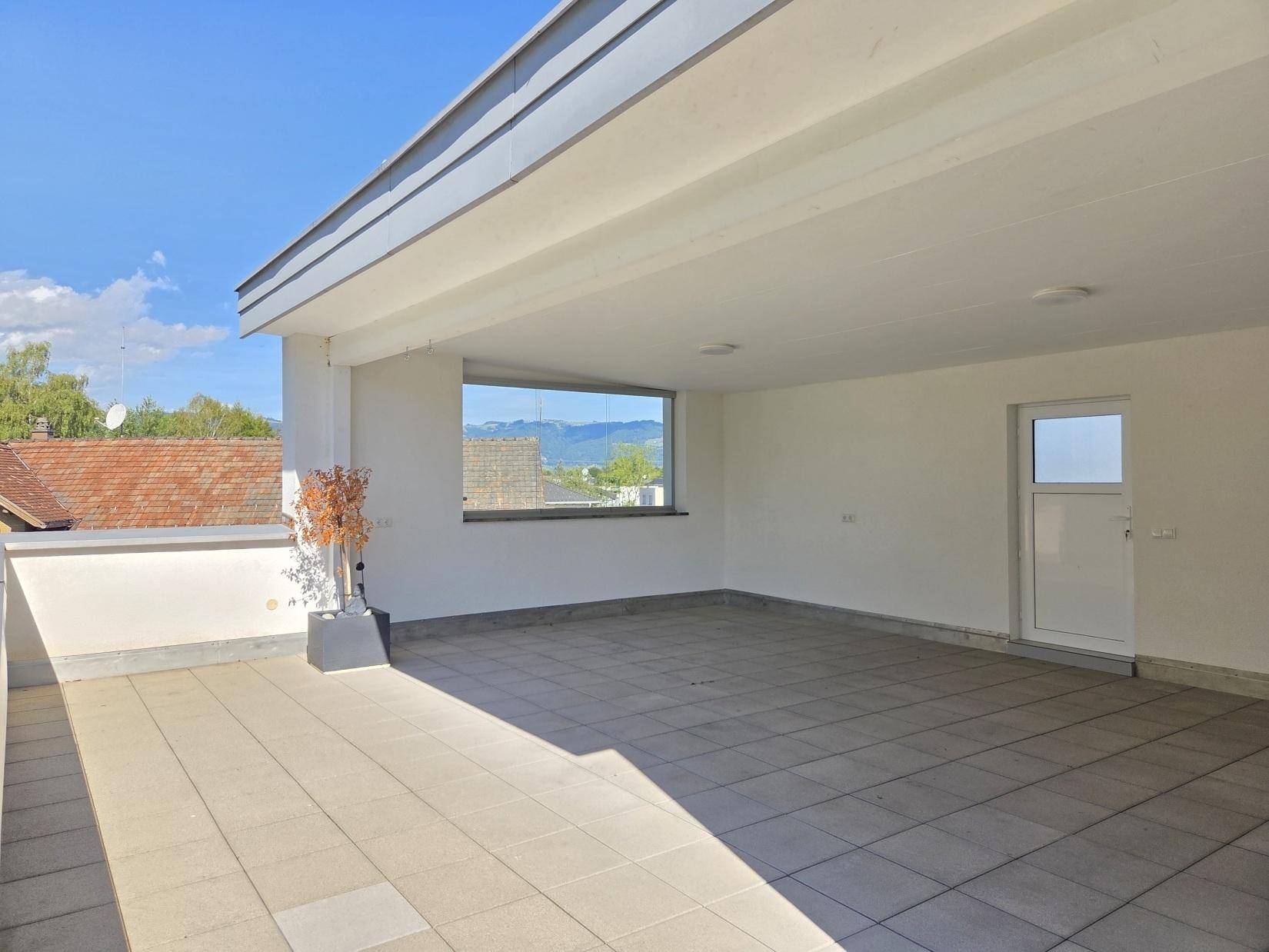 Große, überdachte Terrasse mit Fliesenboden und Fenster, das einen weiten Blick auf die Berge freigibt.