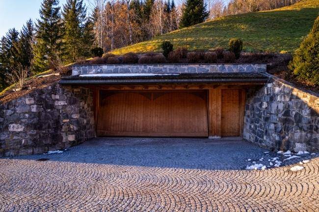 Rustikale Doppelgarage, in den Hang gebaut, mit Holztoren und gepflasterter Zufahrt.