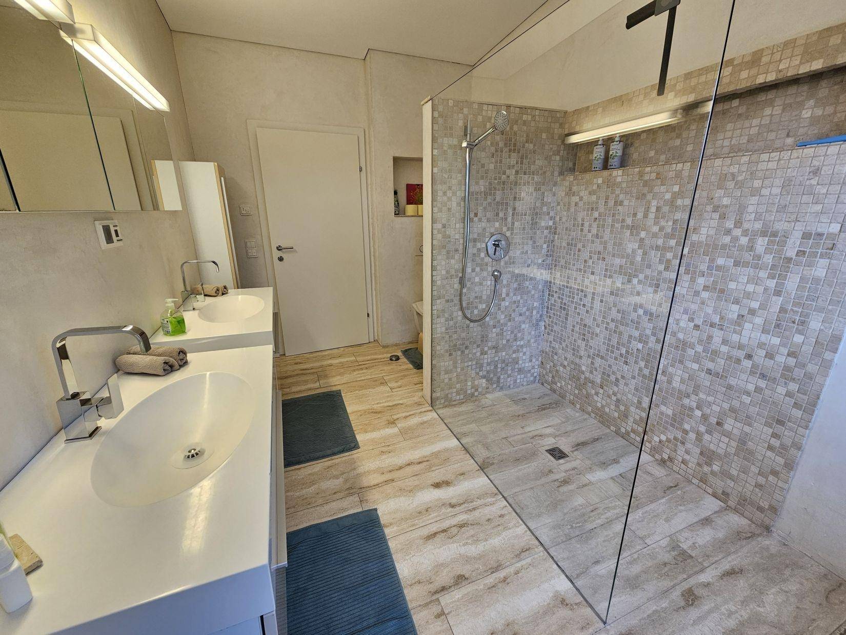 Modernes Badezimmer mit Doppelwaschbecken und großer begehbarer Dusche, hell gefliest.