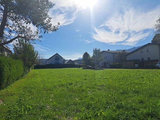 Sonnige Grünfläche mit Hecke, umgeben von Wohnhäusern und Bergpanorama im Hintergrund.