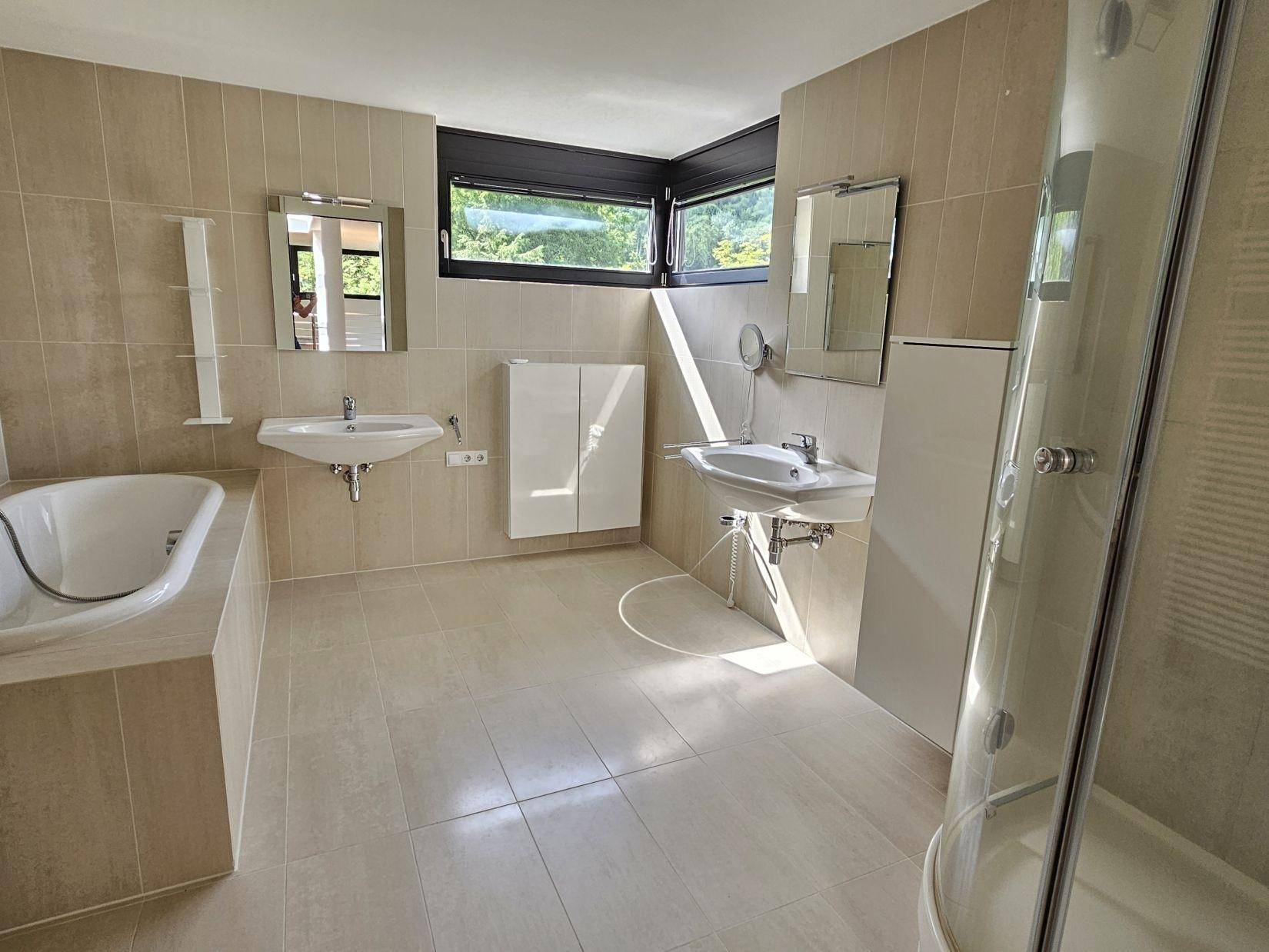Helles Badezimmer mit beigefarbenen Fliesen, Badewanne, Dusche und zwei Waschbecken.