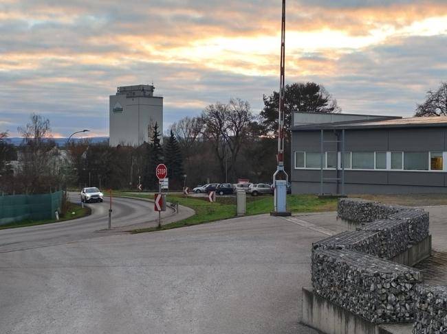 Zufahrt zum Gelände mit Schranke, umgeben von Bäumen und einem Industriegebäude im Hintergrund.