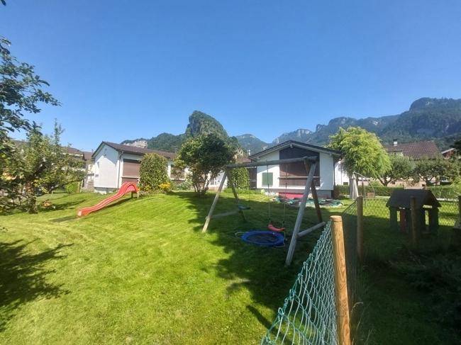 Sonniger Garten mit Schaukel, Rutsche und Spielhaus, umgeben von grüner Natur und Bergen.