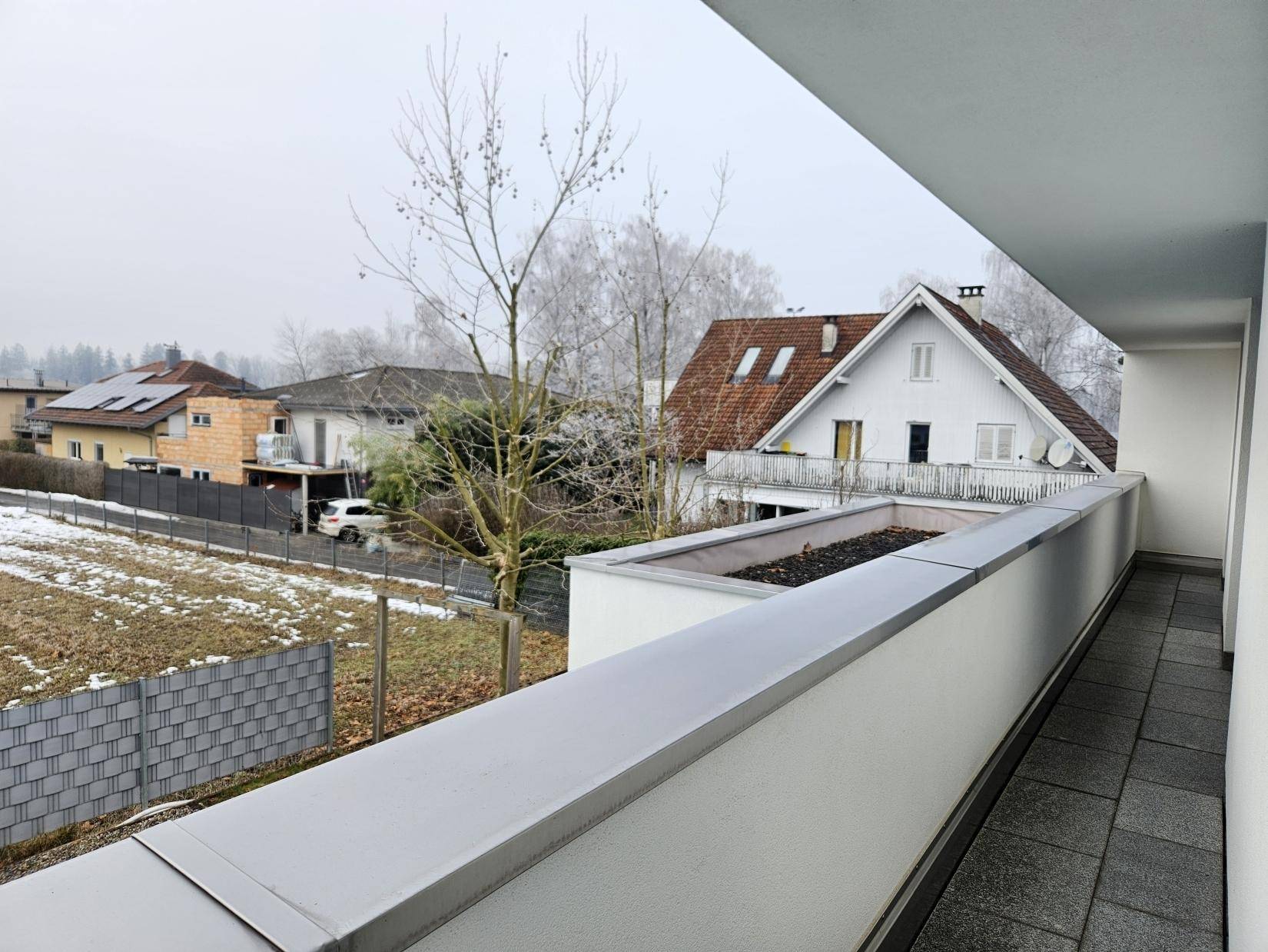 Langer Balkon mit grauen Fliesen und Blick auf die verschneite Umgebung und Nachbarhäuser.