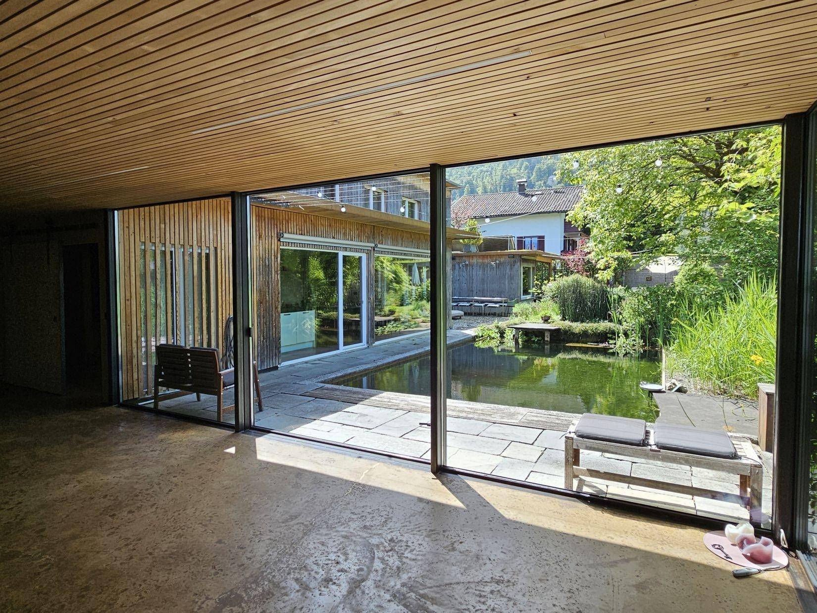 Heller Wohnraum mit großen Glasfronten und Blick auf den Naturschwimmteich und die Außenanlage.