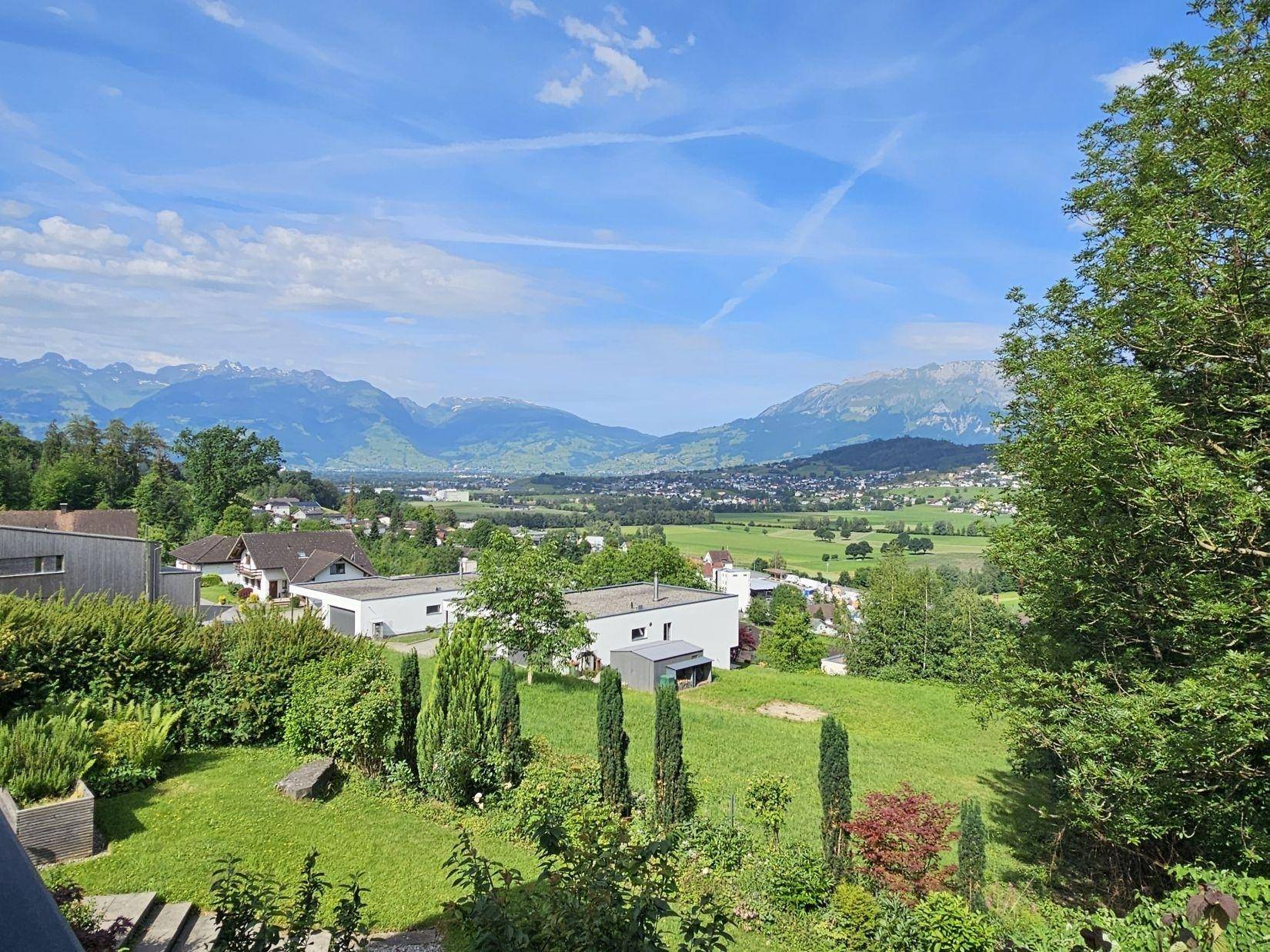 Weitläufiger Ausblick auf die Wohnsiedlung, grüne Felder und die majestätische Bergkulisse.