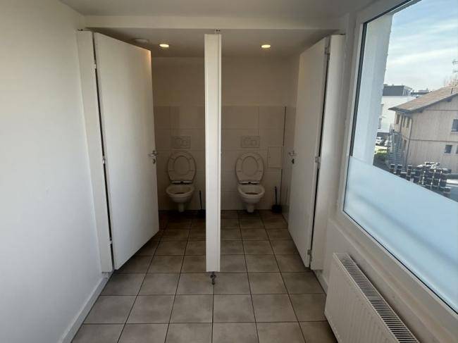 Toilettenbereich mit zwei Kabinen, weißen Fliesen und einem Fenster für Tageslicht.