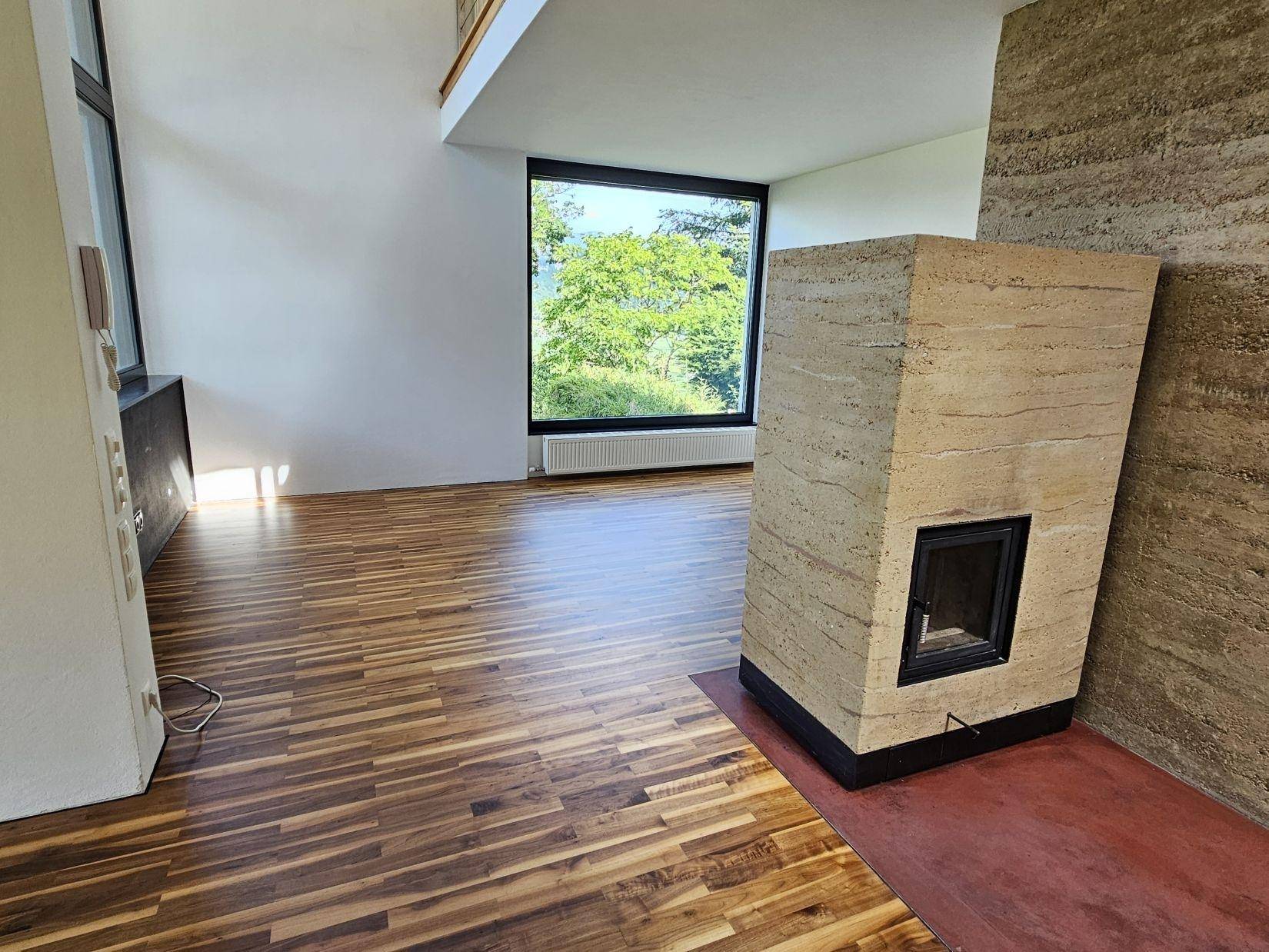 Heller Wohnbereich mit Kamin und großen Fenstern, die einen weiten Blick in die Natur ermöglichen.