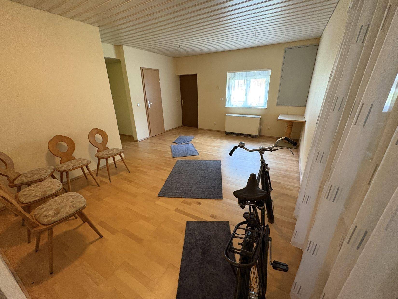Heller Raum mit Holzfußboden, einem Fenster und einer Reihe von Holzstühlen, sowie einem Fahrrad.